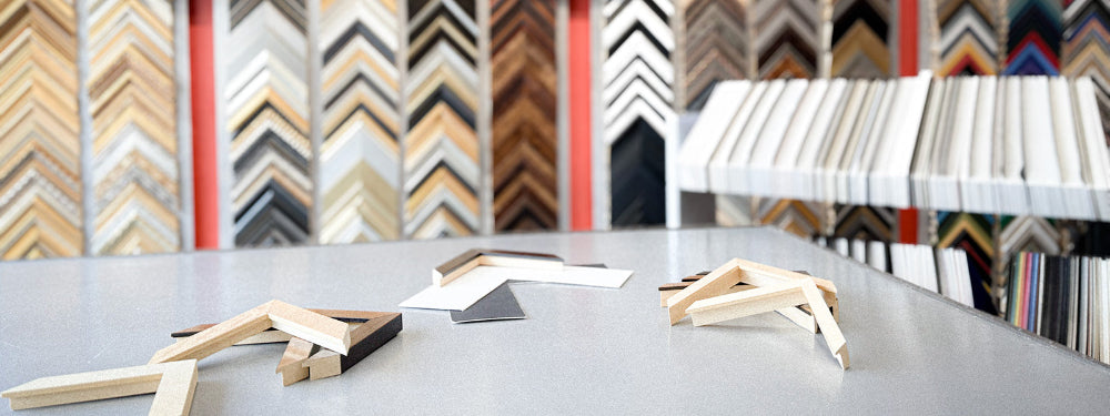 Display of various picture frames on a table with a wall of frame samples in the background.