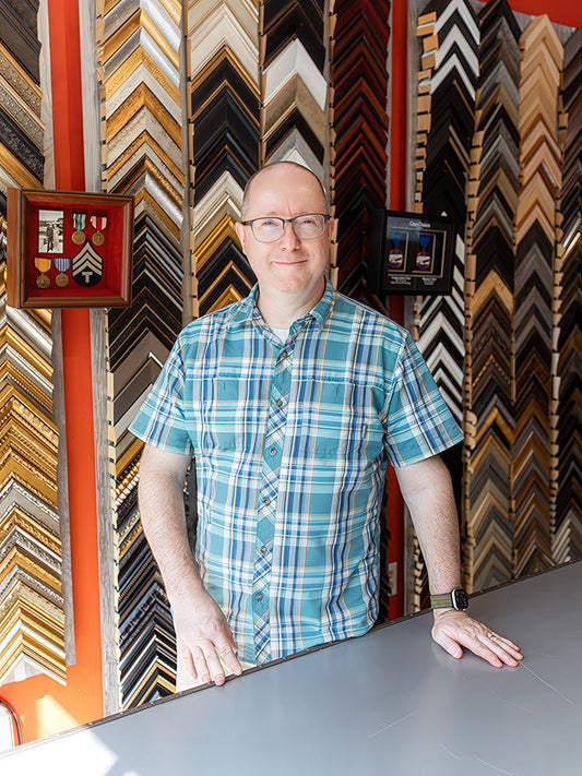 Local custom picture framer standing in his studio, surrounded by frame samples and completed projects.